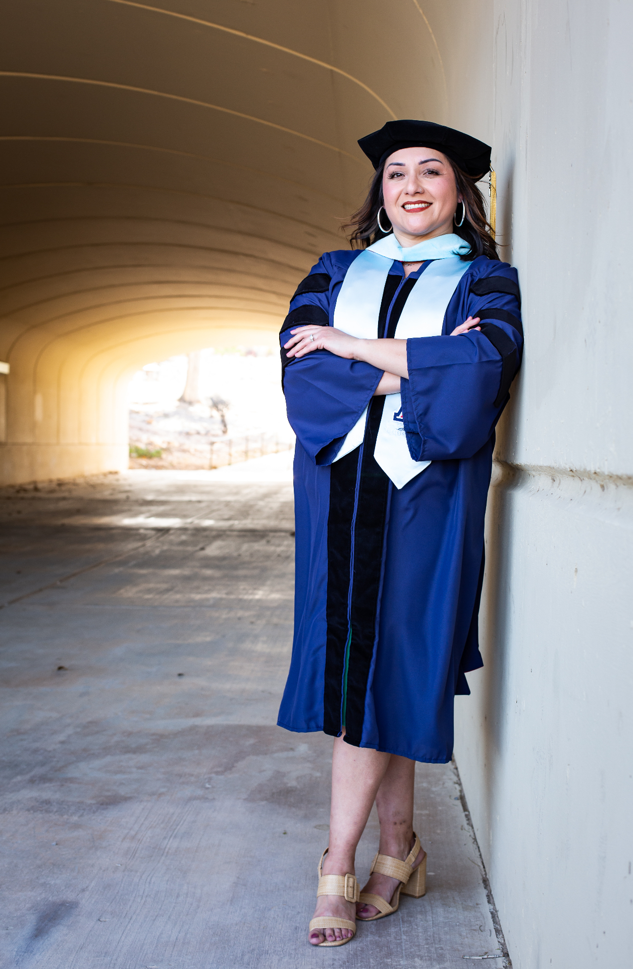 Dr. Martha A. Castillo standing in doctoral regalia, arms crossed, in a light-filled tunnel